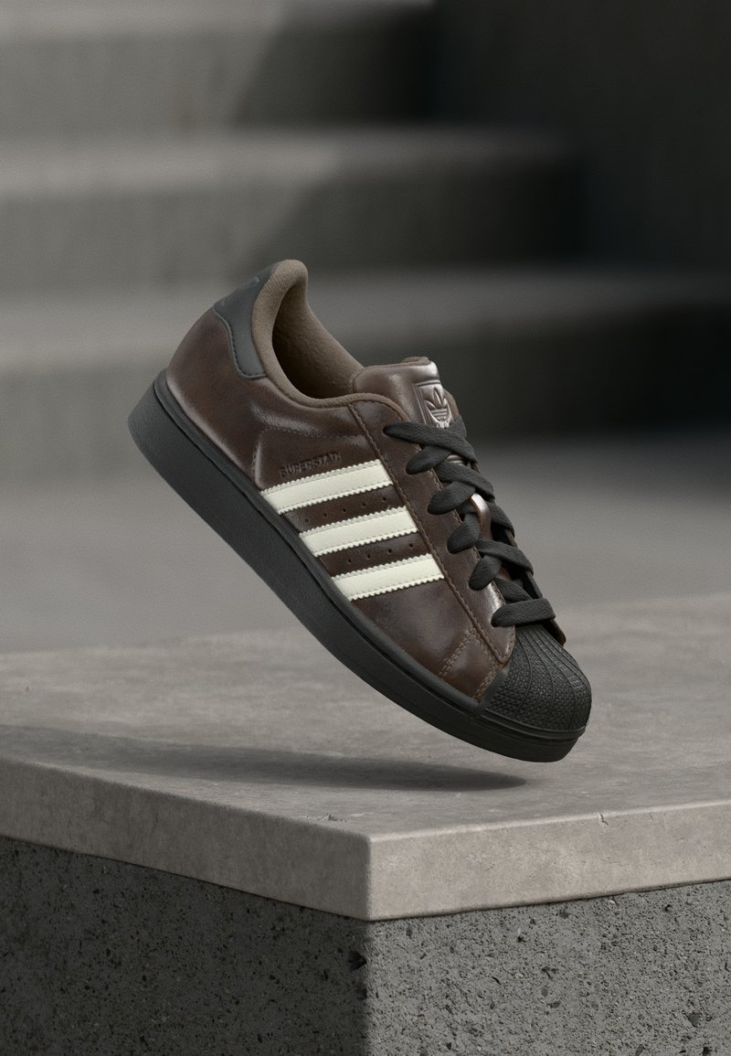 Baskets en cuir marron avec semelle en caoutchouc noire, motif de trois bandes blanches, bout texturé et lacets noirs, sur fond gris.