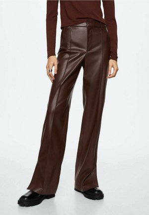 Kobieta ubrana w szerokie spodnie z ciemnobrązowej sztucznej skóry, czarne masywne buty i dopasowaną brązową bluzkę z długim rękawem, stojąca na białym tle.