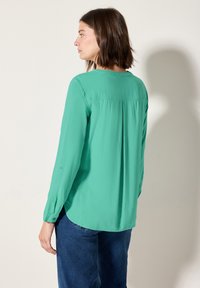 Blouse turquoise à manches longues avec une pliure dans le dos, des poignets boutonnés et un ourlet arrondi, associée à un jean bleu. Texture de tissu lisse.