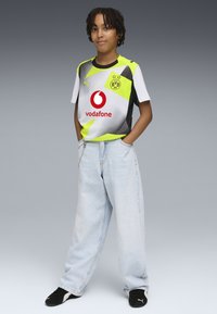 Jersey de manga corta gris y verde neón con acentos negros, que presenta un logo y patrocinadores. Llevado con unos jeans holgados de color azul claro y zapatos negros.
