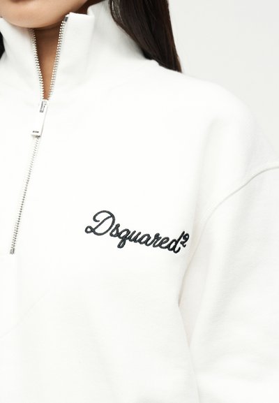 Haut blanc à fermeture éclair avec col montant et logo "Dsquared²" noir brodé sur la poitrine gauche, porté par une personne aux cheveux foncés.