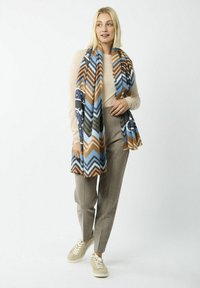 Écharpe à motifs en chevrons multicolores dans les tons de bleu, marron et vert, en tissu doux, tombant sur un haut clair et un pantalon gris.