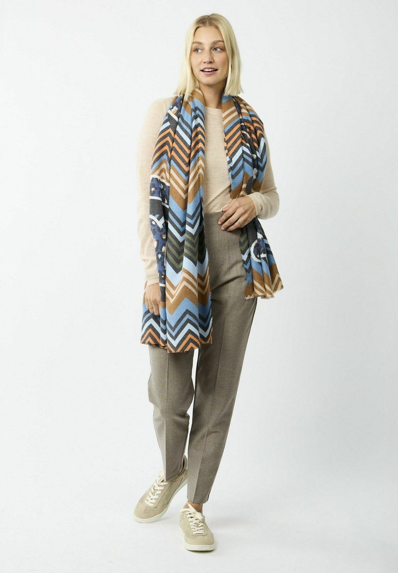 Écharpe à motifs en chevrons multicolores dans les tons de bleu, marron et vert, en tissu doux, tombant sur un haut clair et un pantalon gris.
