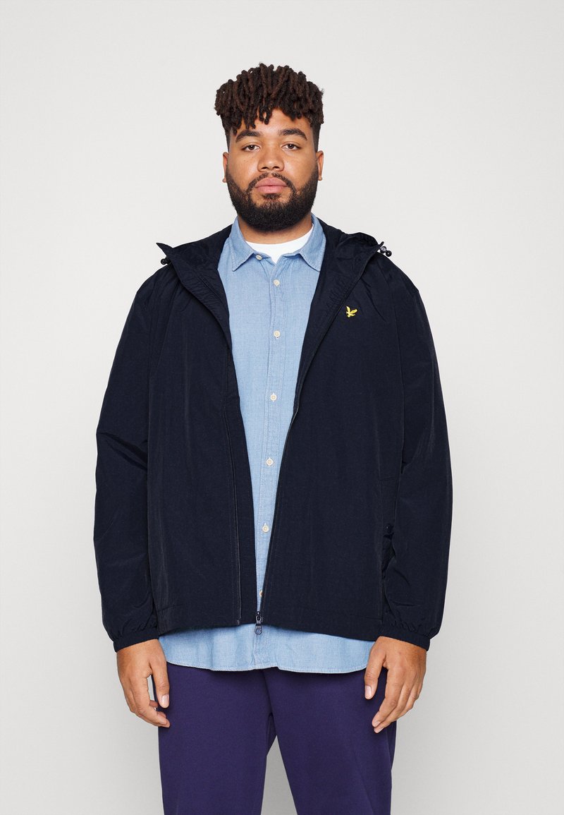 Chaqueta cortaviento navy con capucha y puños elásticos, con cremallera en la parte delantera y un pequeño detalle de logo. Llevada sobre una camisa de botones azul claro.