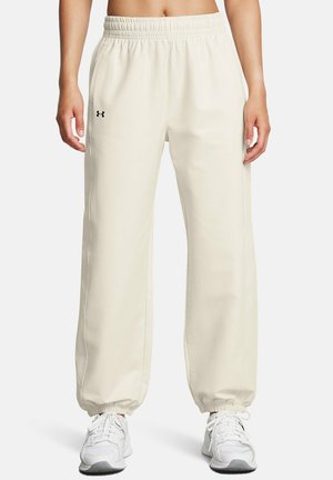 Beige Under Armour Jogginghose mit elastischem Bund und Bündchen, getragen mit weißen Sneakers, von der Taille bis zu den Füßen vor neutralem Hintergrund gezeigt.