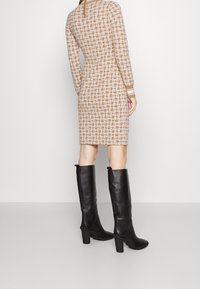 Robe marron ajustée longueur genou avec un motif géométrique crème et des manches longues, associée à de grandes bottes en cuir noires à talons.