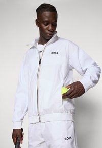 White athletic jacket with a zippered front, ribbed cuffs, and "BOSS" branding. Model holds a tennis ball in the pocket.