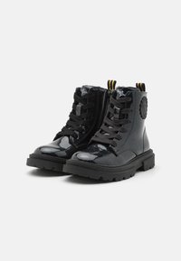 Bottines en cuir verni noir avec des lacets noirs, semelle texturée, tirette jaune et noire, et un accent logo rond sur le côté.