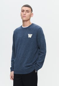 Tmavomodrý pletený sveter s okrúhlym výstrihom, rebrovanými manžetami a lemom, so bielym logom "W" na ľavej hrudi. Mäkká textúra, pohodlný strih.