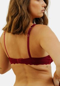Soutien-gorge en dentelle bordeaux avec bretelles réglables et fermeture à crochets. Présente des motifs floraux et des bords festonnés, mettant en valeur une texture lisse.