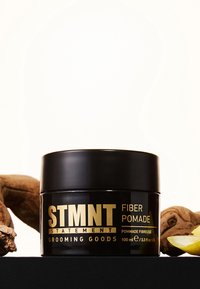 Svart burk med STMT Fiber Pomade, 100 ml, med guldfärgad text. Omgiven av en bit ingefära och en citronskiva på en svart yta.