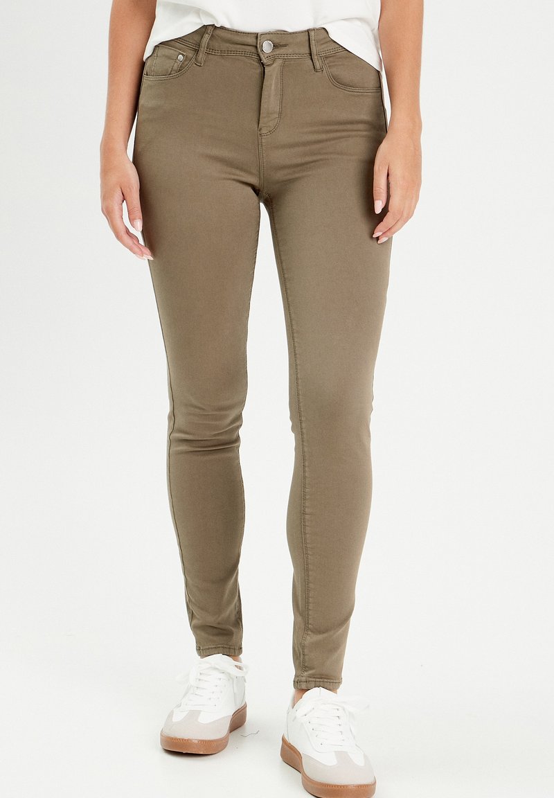 Jeans skinny vert olive à taille haute, avec poches avant et passants de ceinture ; associés à des baskets blanches avec une touche beige.