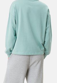 Sudadera de algodón verde claro con mangas largas, corte relajado y escote redondeado, combinada con pantalones de chándal gris claro con bolsillos.