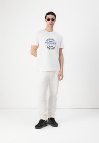 White cotton t-shirt with blue and red "Original 1985" graphic, paired with white pants and black sneakers, standing against a white background.