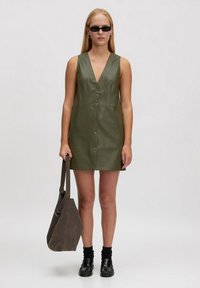 Olivenfarbendes ärmelloses Kleid mit tiefem V-Ausschnitt, Knopfdetails über die Vorderseite und glatter Textur. Kombiniert mit einer dunklen Tasche und schwarzen Schuhen.