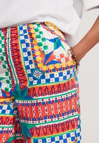 Pantalon à motifs multicolores avec des designs géométriques en tissu rouge, bleu, vert et jaune. Taille élastique, détail visible de poche.