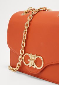 Borsa a tracolla in pelle arancione con catena dorata, dotata di una chiusura dorata e una texture liscia. Design rettangolare compatto.