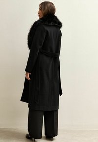 Cappotto nero in lana con colletto in pelliccia sintetica, maniche lunghe e cintura. Presenta una vestibilità rilassata, tasche laterali e un design non strutturato.