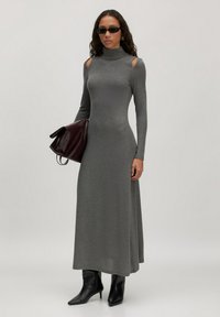 Femme portant une robe longue grise ajustée avec des découpes aux épaules, des bottes noires, de petites lunettes de soleil noires, tenant un grand sac brun foncé.