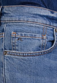 Ljusblå denimjeans med orange sömmar, en liten silverlogotyp på bakfickan, och en klassisk femficksdesign.