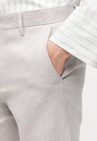 Άτομο που φοράει ανοιχτό μπεζ παντελόνι με ψαροκόκαλο και το χέρι στην τσέπη, και λευκό πουκάμισο με λεπτές γκρι κάθετες ρίγες.