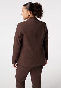 Bruine op maat gemaakte blazer met een gestructureerd ontwerp, voorzien van drie knopen op de manchetten en een gladde, getextureerde stof. Achteraanzicht weergegeven.