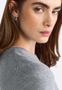 Suéter de punto gris con escote redondo. Presenta una textura suave y un sutil detalle de costura, usado por un modelo con cabello largo y liso.