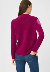 Magenta Pullover mit lockerer Passform, langen Ärmeln und einem kontrastierenden pinken Patch auf der Schulter, kombiniert mit blauen Jeans. Weicher Stoff und abgerundeter Saum.