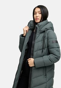 Cappotto invernale imbottito verde con colletto alto, chiusura a zip e cappuccio con cordino. Presenta un patch con logo sulla manica e un motivo a trapuntatura.