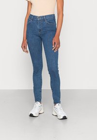 Calças skinny de denim em azul médio, de cintura alta com bolsos frontais. Combinadas com sapatilhas brancas e uma blusa às riscas, que apresenta um ajuste justo.