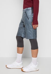 Blå isolerade shorts med elastisk midja, sidofickor med dragkedja, i kombination med grå caprileggings och vita sportskor.