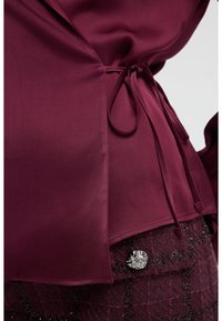 Bordeaux satinbluse med en bindebånd i taljen, der har en glat overflade og en buet nederkant, parret med mørkt struktureret stof og en sølvknap.