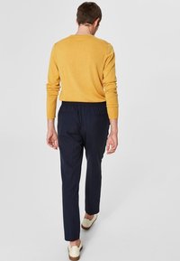 Geel gebreide trui gecombineerd met marineblauwe elastische broek en lichte schoenen, gezien vanuit de achterkant. De textuur lijkt zacht en ontspannen.