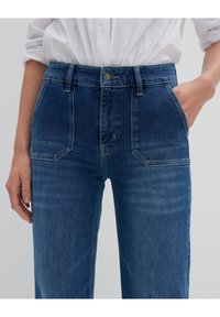 Jeans alta vita in denim blu, con tasche anteriori, cuciture laterali e un bottone di chiusura in vita, dalla finitura liscia e leggermente strutturata.