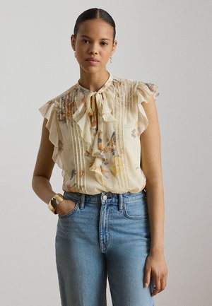 Femme aux cheveux tirés en arrière portant un chemisier léger à volants à motif floral noué au cou, un jean bleu et un bracelet manchette doré au poignet gauche.