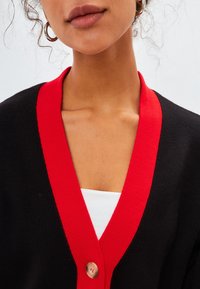 Black cardigan with a red ribbed collar and a button detail at the chest, worn over a white top, highlighting contrasting colors and textures.