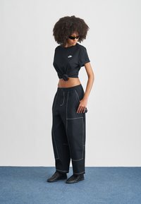 Νεαρή γυναίκα με σγουρά μαλλιά που φοράει μαύρα γυαλιά ηλίου, κοντοκομμένο μαύρο μπλουζάκι Nike, φαρδιά μαύρη παντελόνα με λευκές ραφές και μαύρα παπούτσια.