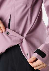 Ljuslila utomhusjacka med tryckknappsstängning, sidoficka och svarta kardborre-accenter vid ärmslutet för justerbar passform.