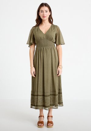 Woman wearing olive green midi dress with flutter sleeves and small floral pattern, paired with brown platform sandals, standing.