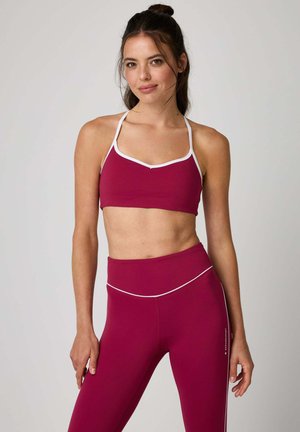 Woman with dark hair in a bun wearing a berry-colored sports bra and matching high-waisted leggings with white trim, standing against a plain background.