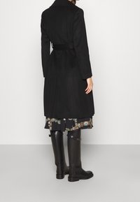 Svart lång kappa med bälte i midjan, med notchkrage och slät textur, kombinerad med svarta knähöga stövlar och en mönstrad klänning.