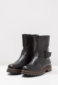 Botas de tobillo de cuero negro con cierre de cremallera, detalle de correa de cinturón, suela texturizada robusta y tacón ligero. Acabado suave con acentos de costura.