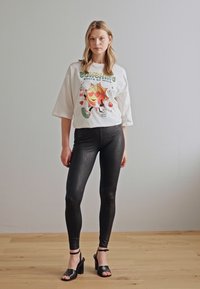 T-shirt blanc oversize avec un graphique de soleil coloré, associé à des leggings noirs brillants avec un motif texturé et des sandales à talons noires.