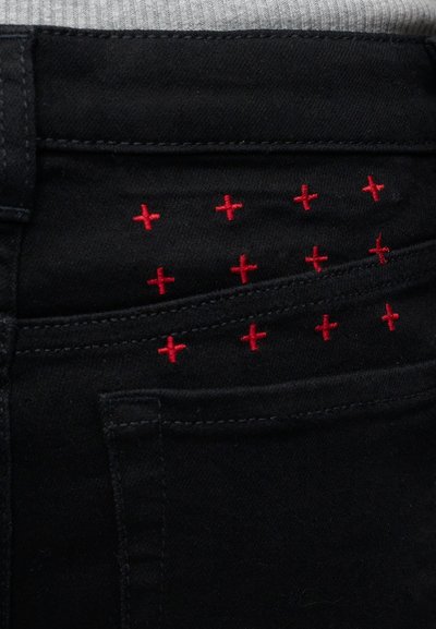 Μαύρη τσέπη από τζιν με κόκκινα ραφές σε σχήμα σταυρού πάνω από τη ραφή και το θηλάκι της ζώνης σε φόντο γκρι υφάσματος.