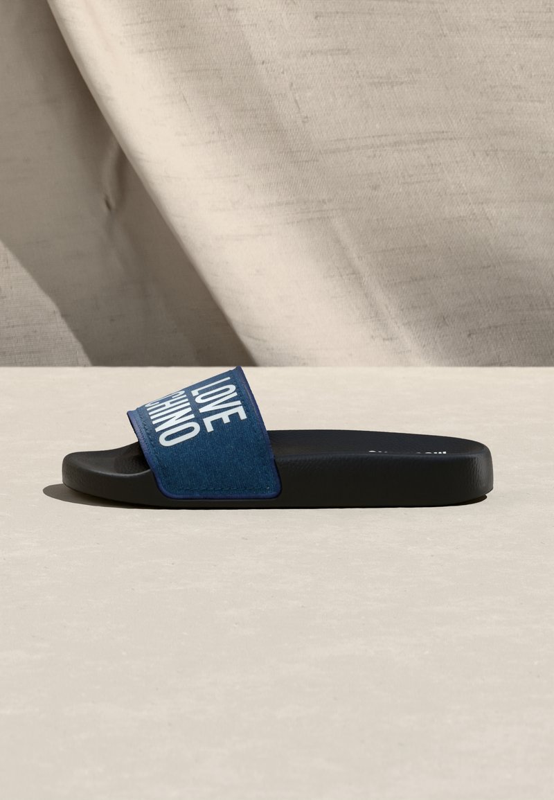 Sandale slide noire avec une bride en tissu bleu marine ornée du texte blanc en gras "LOVE MOSCHINO", sur un fond texturé beige.