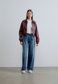 Bordeauxrood leren cropped jack over een wit shirt, gecombineerd met lichtblauwe wijde jeans en witte sneakers op een neutrale achtergrond.