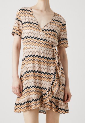 Kobieta ubrana w sukienkę kopertową z krótkim rękawem o zygzakowatym wzorze w odcieniach brązu, czerni i beżu, z falbanianym dołem i dekoltem w kształcie litery V.