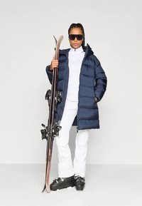 Kvinna i en mörkblå puffjacka, vit skidoutfit, svarta skidstövlar och solglasögon, som håller ett par skidor upprätt bredvid sig.