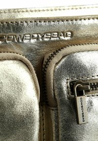 Borsa in pelle metallica dorata con due tasche con zip. Superficie testurizzata, accenti in tessuto beige a contrasto e dettaglio della zip con marchio.