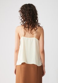 Camicetta in seta color crema con spalline sottili, caratterizzata da una vestibilità leggermente ampia e un orlo curvo, abbinata a una gonna marrone fluida.